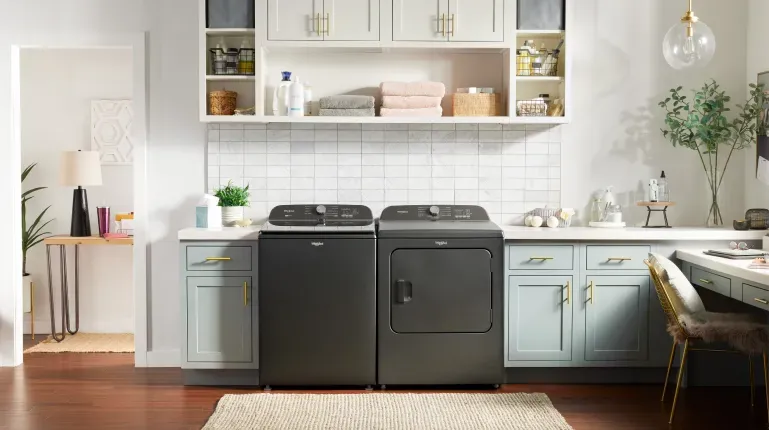 Whirlpool WTW6157PB High-Efficiency Top-Load Washer and matching dryer in dark chrome finish, set in a bright laundry room with blue cabinets